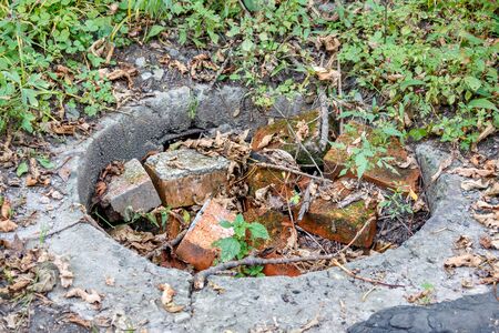 The concrete well of the city drainage system is overgrown with grass and filled with stones and mud with branches. An abandoned storm drains sump is not serviced and has problemsの写真素材