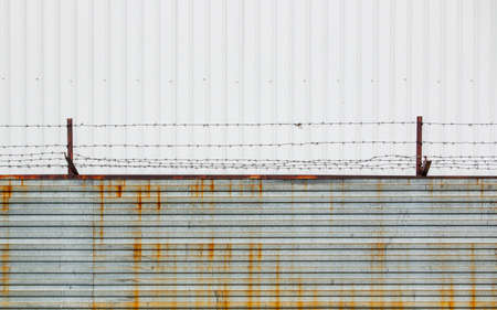 Barbed wire stretched over rusty brackets mounted on a fence made of metal sheets against a cloudy...