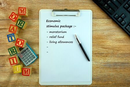 Conceptual of moratorium by banks as one of economic stimulus packages during crisis. Top view keyboard, calculator, pen, texts on white paper, alphabet cubes. Focus on top of clip board.の写真素材
