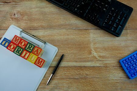 Top view of conceptual of moratorium by banks during financial crisis and recession. Keyboard, calculator, pen, white paper, alphabet cubes on MORATORIUM on wooden table. Focus on top of clip board.の写真素材