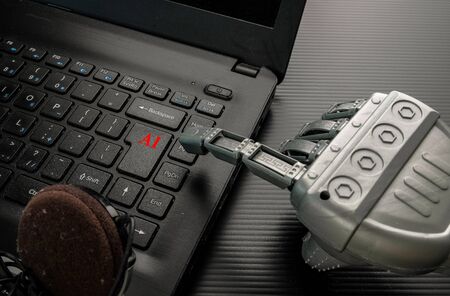 Conceptual image of artificial intelligence (AI). Robotic hand pointing to keypad with red texts of a black notebook. Earphone partly visible. Selective focus on keypad with red texts.の写真素材