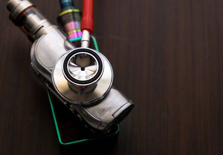 Conceptual image of uncertainty of health safety of e-cigarettes. Isolated close-up red stethoscope diagnosing  vaping kits over dark background. Focus on bell-diaphragm part.の写真素材