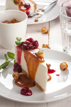 Naked cheesecake slice topped with sour cherries and caramel sauces and caramelized hazelnuts served on on white plate over white table, close upの写真素材
