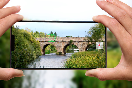 Woman with mobile phone photos old stone bridge.