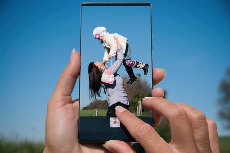 Woman photographing on cell phone cute young daughter playing in nature with her mother.の写真素材