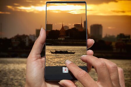 Woman photographing on cell phone sunset on the river with boat and temple wat in the background in Thailand.の写真素材