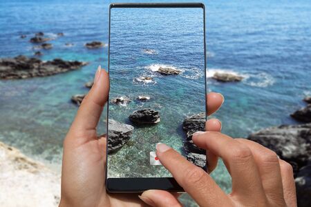Woman photographing on cell phone rocky beach from cliff.