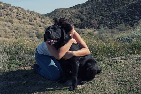 Woman sitting on ground cuddle and kiss Labrador dog while walking in nature.