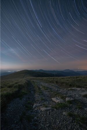 Star trails in the night sky above the road leading through the mountain ridge