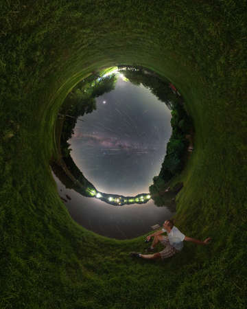 360 degree Panorama of the night sky with the Milky Way and the meteor shower Swarm 2021. A man sits on a meadow by the lake.の写真素材