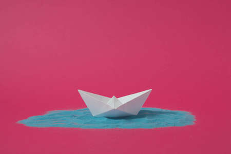White origami paper boat floating on blue sand with pink background. Minimal concept.の写真素材