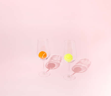 A yellow and an orange tennis ball in two transparent wine glasses on a pink backgrouns. Minimal concept.の写真素材