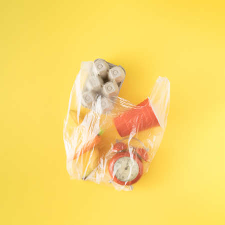 Plastic transparent bag with cardboard egg box, carrot, banana, clock and plastic cup on yellow...