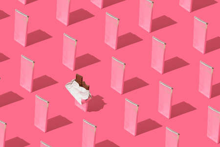 Pattern made of chocolate bars wrapped in aluminum foil with one unpacked bar on pink background.