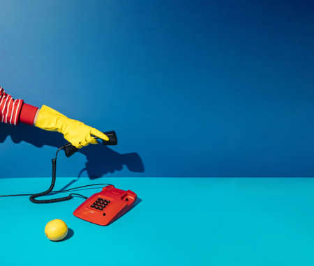 Minimal retro concept with hand in yellow cleaning glove answering red vintage phone on blue background.の写真素材