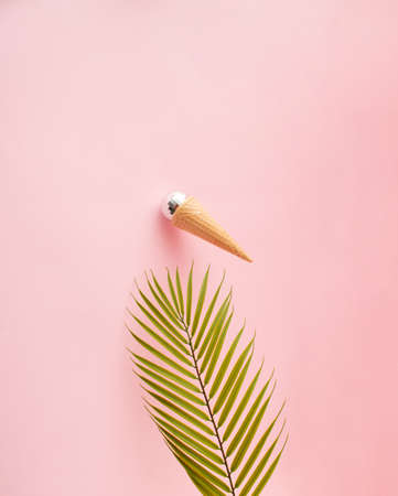 Ice cream cone with shiny disco ball sticking on the pink wall above palm leaf. Front view. Minimal concept.の写真素材