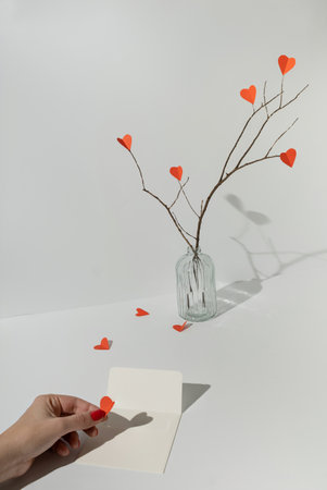 Branches with red paper hearts and female hand putting heart in white envelope. Minimal love or valentine concept.の写真素材