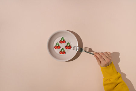 Hand with fork reaching for gummy candies shaped like cherries served in plastic plate on beige...