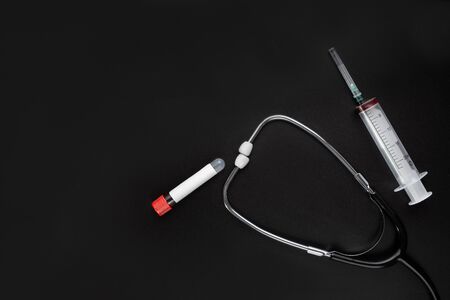 A test tube for taking blood samples for the detection of coronavirus with a blank label for inserting text, a stethoscope and a syringe on a black background. The concept covid-19 or 2019-nCov.の写真素材