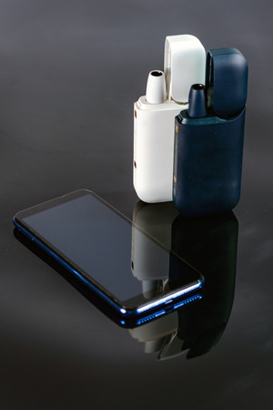 The devices for heating  tobacco cigarettes  with a charger. Charger itâs a case . The smartphone  located next to charge on a black mirror backgroundの写真素材