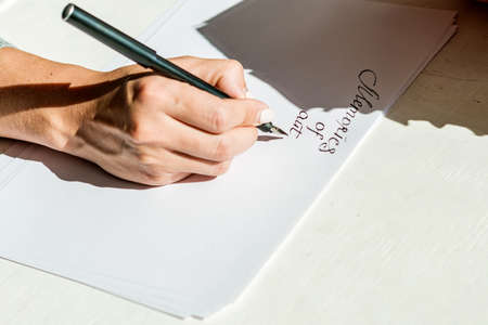 The woman's hand draws a close-up of English letters on a white sheet of paper. Calligraphic inscriptions by women's hands with a fountain pen under a bright light.の写真素材