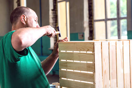 A man in a green t-shirt, hammering nails into a wooden box. The concept of working on wood products.の写真素材
