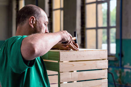 A man in a green t-shirt, hammering nails into a wooden box. The concept of working on wood products.の写真素材