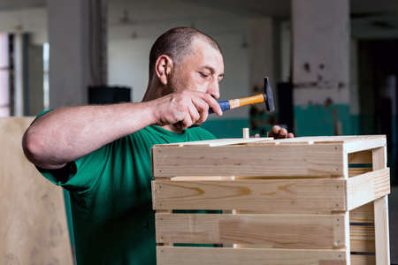 A man in a green t-shirt, hammering nails into a wooden box. The concept of working on wood...