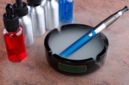 Electronic cigarette on glass ashtray and vape liquids within vapor on granite surface.