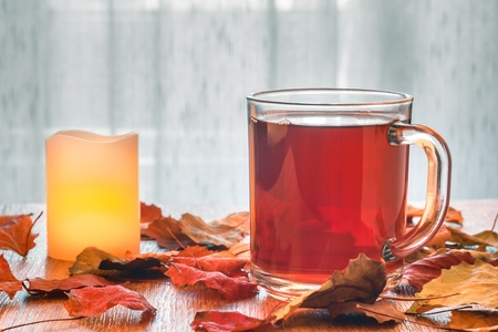 Glass cup of tea and glowing candle on wooden table with autumn leaves. Light tulle backgroundの写真素材