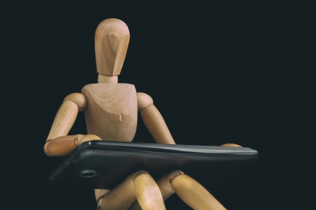 Sitting wooden dummy works with smartphone that lies in his lap. Dark backgroundの写真素材