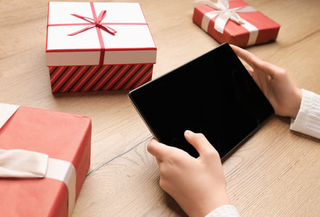 Hands hold a tablet on a wooden background, boxes with gifts. Ordering gifts via the Internet. Internet - shopping, sale, black friday. Christmas spirit, holiday concept, surprises for children. Gifts for the New Year. The gadget is in children's hands.の写真素材
