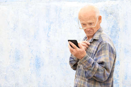 Grandpa with gray hair holds a phone in his hand against a brick wall. A man with glasses makes a phone call. Pensioner on a light background. Old age and new technologies.の写真素材
