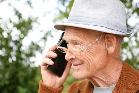 Mature man with gray hair holds a phone in his hand on the nature background A man in glasses makes a phone call. Pensioner on a green background. Old age and new technologies.の写真素材