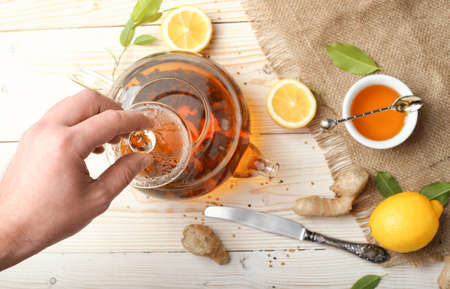 A man's hand holds the lid of a glass teapot with tea. Ginger, half a lemon and honey in a bowl on a burlap top view. Useful vitamin ingredients on a light wooden background.の写真素材