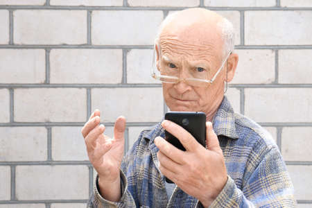 Grandpa with gray hair holds a phone in his hand against a brick wall. A man with glasses makes a phone call. Pensioner on a light background. Old age and new technologies.の写真素材