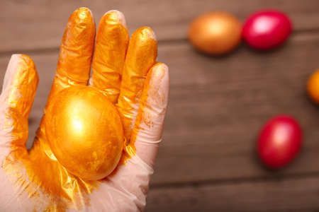 Female hand in a glove holds a painted golden egg on a wooden table close-up. Preparing for the holiday of bright Easter. Art of coloring, decor for Easter eggs.の写真素材