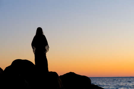 Silhouette of a woman on a rocky beach at sunsetの写真素材