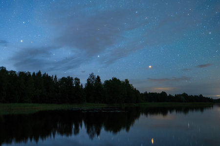 Night landscape under starry sky. Gemini and Orion constellations rising above horizon, stars reflecting from calm water surface.の写真素材