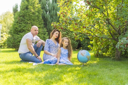 Dad, mom and daughter are sitting on the grass in the park. The whole family is happy together. Her daughter has a globe, she studies at school.の写真素材