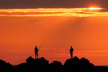 Silhouette of two fishermen fishing at sunset on the rocks.の写真素材