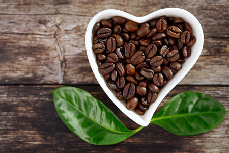 top view of roasted coffee beans in white heart-shaped bowl on wooden background with green leaves of coffea arabica, concept for coffee loversの写真素材
