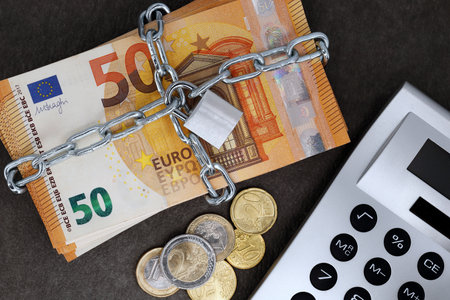 A stack of cash with 50 euro banknotes is locked with a chain and padlock next to a calculator and coins, concept for the safe investment of fixed-term deposits or financial security in the futureの写真素材
