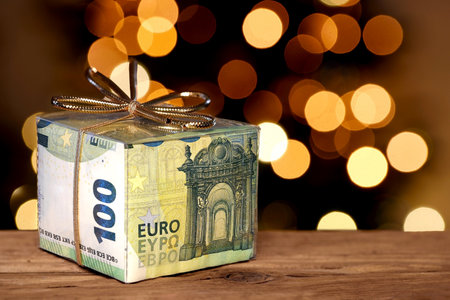 One hundred euro banknotes in transparent gift box with golden bow in front of lights bukeh of a christmas tree on wooden table, Concept of Christmas bonus or giving away moneyの写真素材