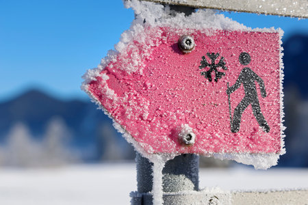 Close-up of a frozen sign showing the hiking trail in the mountains on a sunny, cold winter day, pink signpost covered with ice crystals in a snowy winter landscapeの写真素材