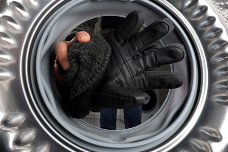 male hand puts black skiing gloves and beanie in the washing maschine, Ski gloves should not be...