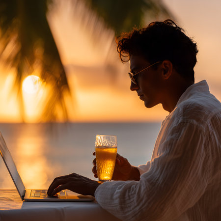 Young man using laptop on the beach at sunset, drinking orange juiceの素材