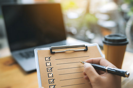 Close up of businesswoman hand writing on clipboard with checklist and laptop computer on wooden table.の素材