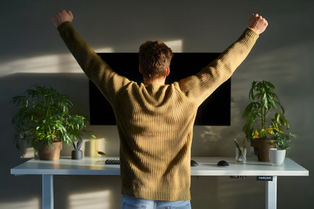 Rear view of young man sitting at the desk and raising his hands up.の素材