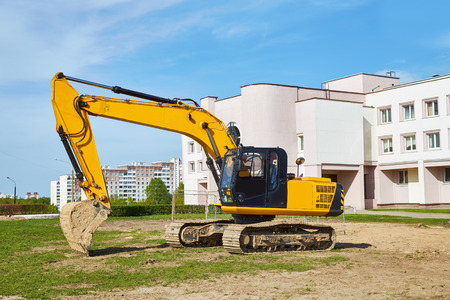A yellow excavator with a black cab stands on the field, in the background residential buildings
